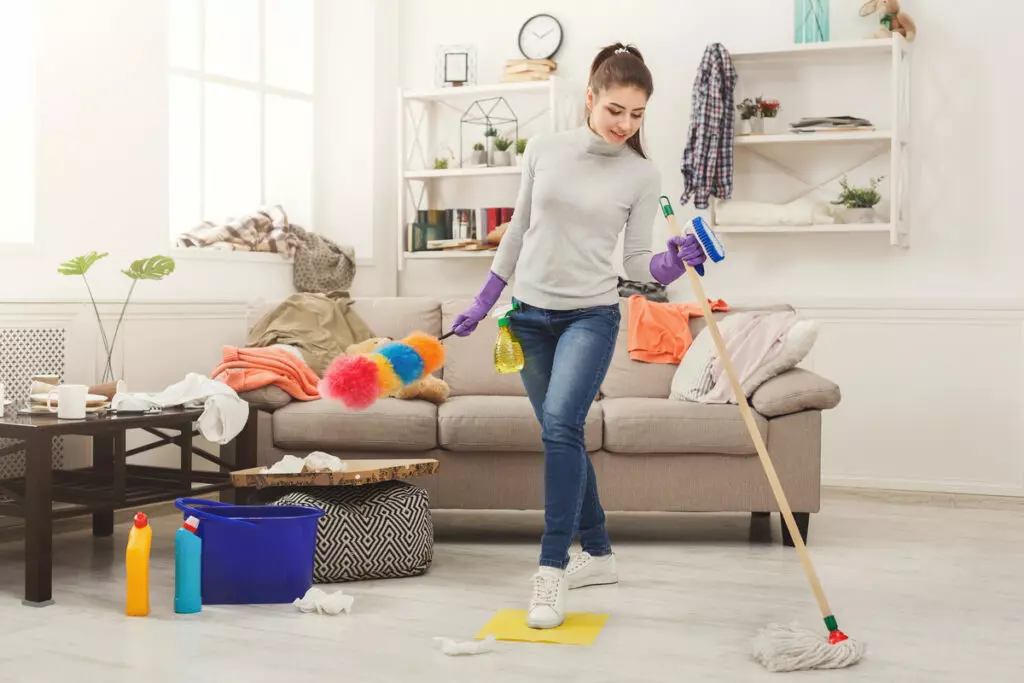 a cleaning lady wearing gloves, holding a mop and a feather duster, with a bucket and disinfectant liquid next to her