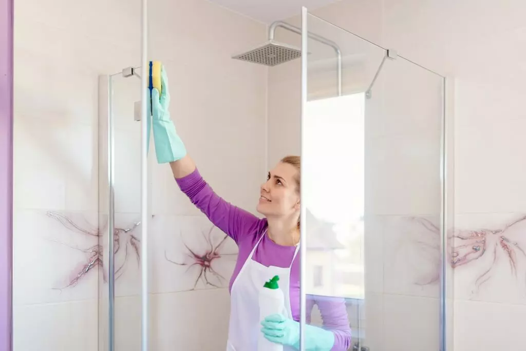 an image of a woman wearing gloves, using a sponge to clean a fiberglass shower with one hand, while holding a bottle of liquid disinfectant in her other hand