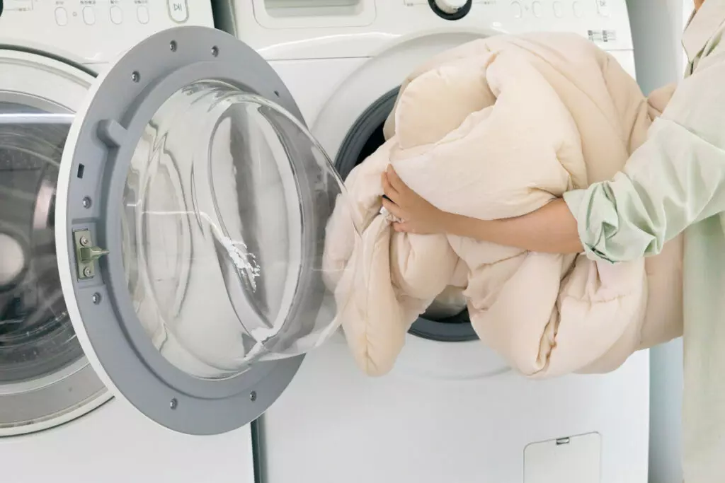an image of a hand carefully placing a blanket into the front loading compartment of a washing machine; how to wash blankets