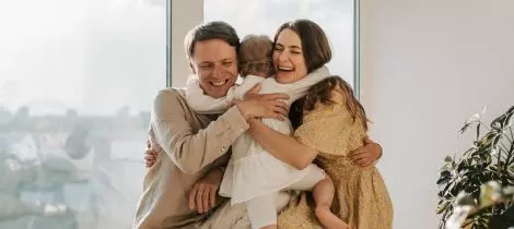 A family is hugging their baby in front of the window.
