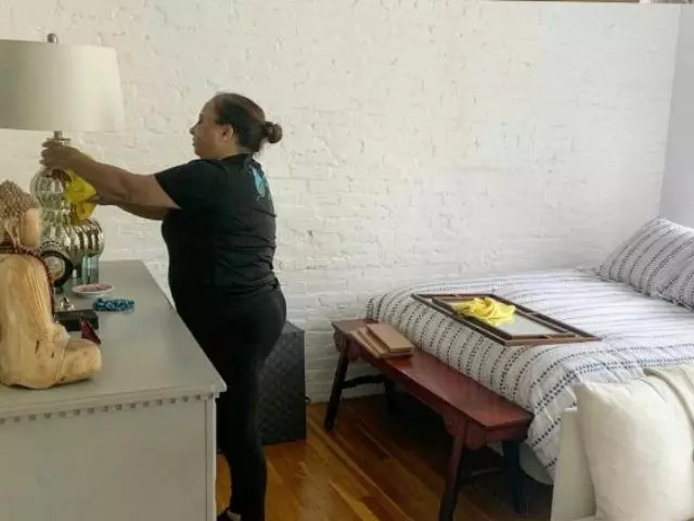 Woman adjusting a lampshade on a bedside table in a bedroom with white brick wall during bedroom cleaning.