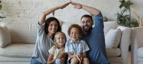 A family of four sitting on a couch, smiling, with the parents forming a roof-like shape with their arms above their children's heads after a day of house cleaning.