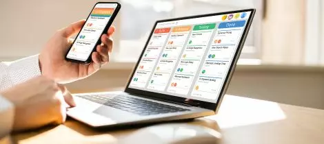 A man is using a laptop and a cell phone at the same time to organize recurring cleaning schedules.