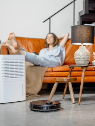 A woman relaxes on a couch with her feet up, a portable air purifier nearby, and a robotic vacuum cleaner moving on the floor in a modern living room, following studio cleaning templates.