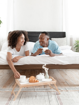 A couple sitting on a bed, enjoying breakfast with coffee and pastries on a tray in a bright studio bedroom.