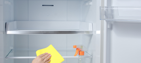 A person's hand wiping the interior of an empty refrigerator with a yellow cloth, with steam cleaning spray visible.
