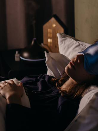Woman sleeping in bed with a blue sleep mask, a humidifier running in the background, and a small lit house-shaped lamp on the nightstand featuring studio cleaning templates.