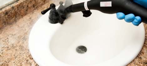 A person in blue gloves steam cleaning a white sink with a black hose attachment.