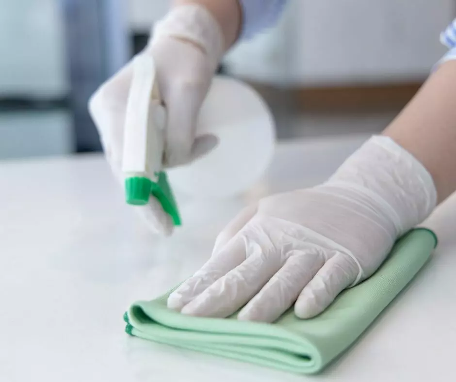 Person wearing gloves performing spring cleaning on a white surface with a green cloth and spray bottle.