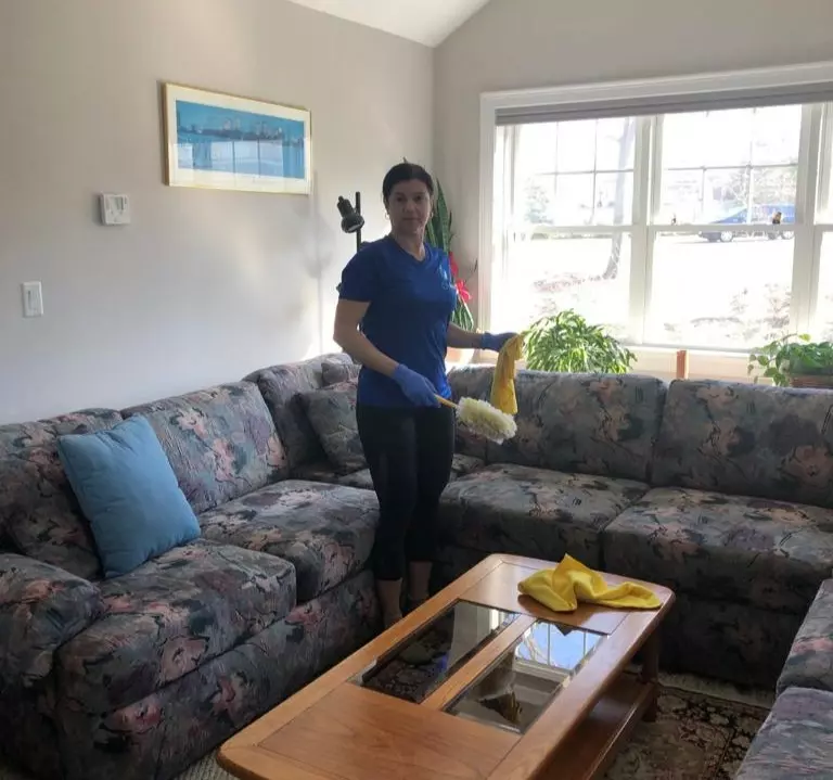 A person standing in a living room holding a dusting cloth, with a couch, coffee table, and two windows in the background, following cleaning templates for efficient living room cleaning.