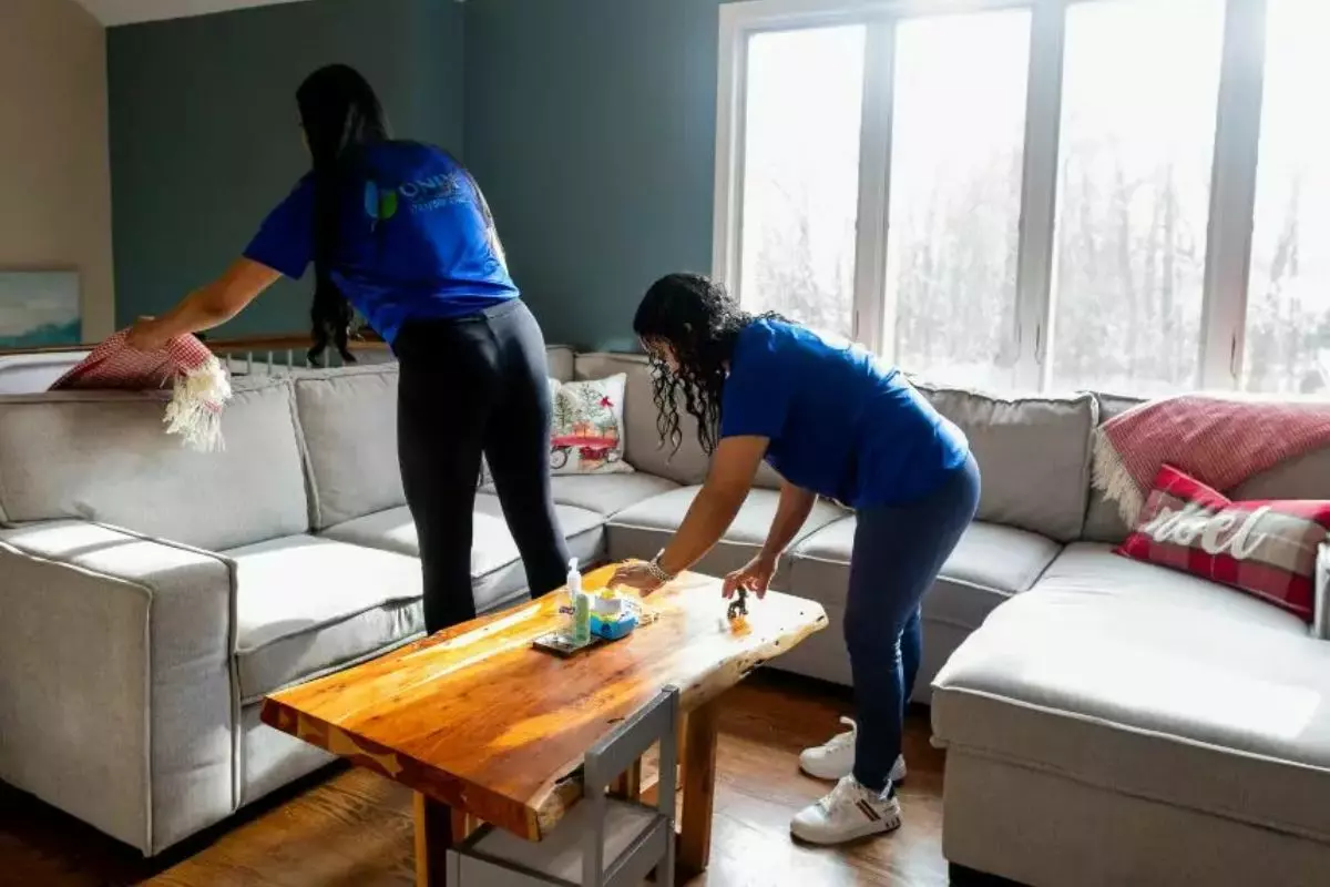 Two individuals wearing blue shirts are tidying a living room. One is arranging a throw blanket on a sofa, while the other is organizing items on a wooden coffee table, showcasing their efficient cleaning services.