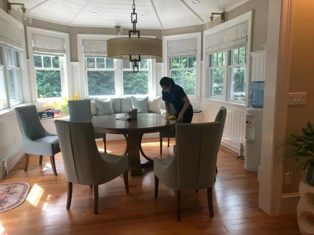 A person is providing cleaning services at a round dining table in a sunlit room with large windows, cushioned seating, and upholstered chairs.