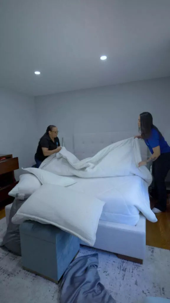 Two people are making a bed in a room with white walls and a gray headboard. They are placing a large white blanket on the bed. Pillows and a blue ottoman are in the foreground, suggesting that expert services were involved in designing the harmonious space.