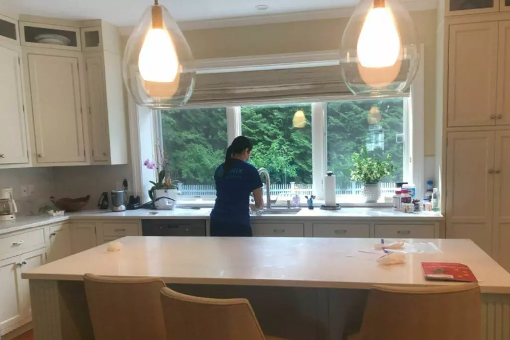 A person is seen from the back washing dishes at a sink in a bright kitchen with white cabinets, large windows, and pendant lights, embodying the meticulous nature of top-tier cleaning services.