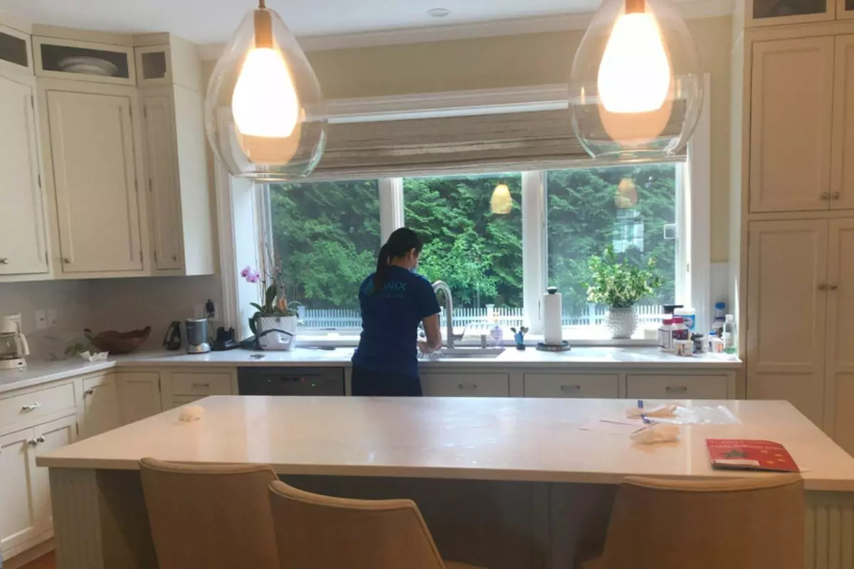 A person is seen from the back washing dishes at a sink in a bright kitchen with white cabinets, large windows, and pendant lights, embodying the meticulous nature of top-tier cleaning services.