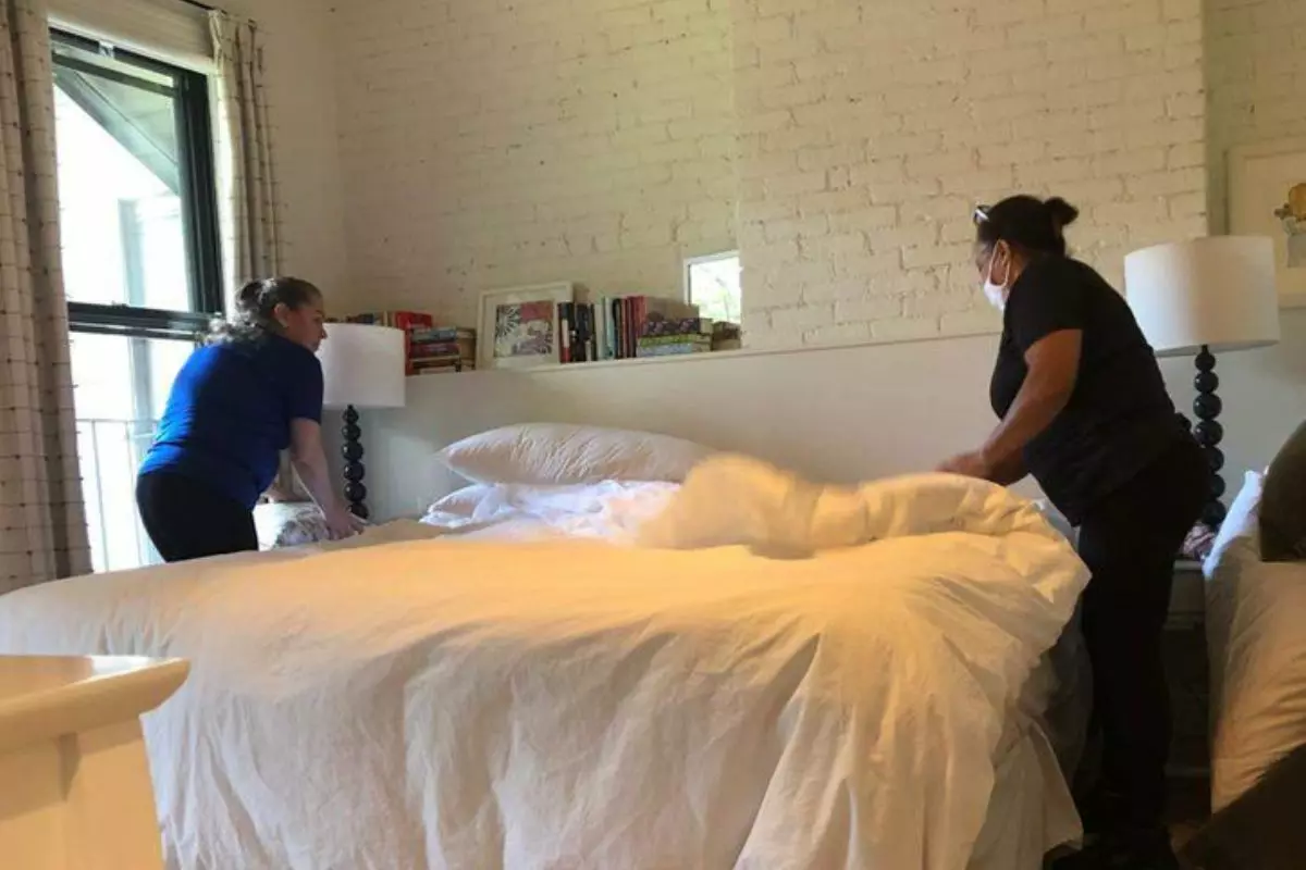 Two people are making a bed in a room with white brick walls and a window. The bed has white sheets, and there are books and lamps on the nightstands, creating an inviting space perfect for enjoying quiet services or personalized care.
