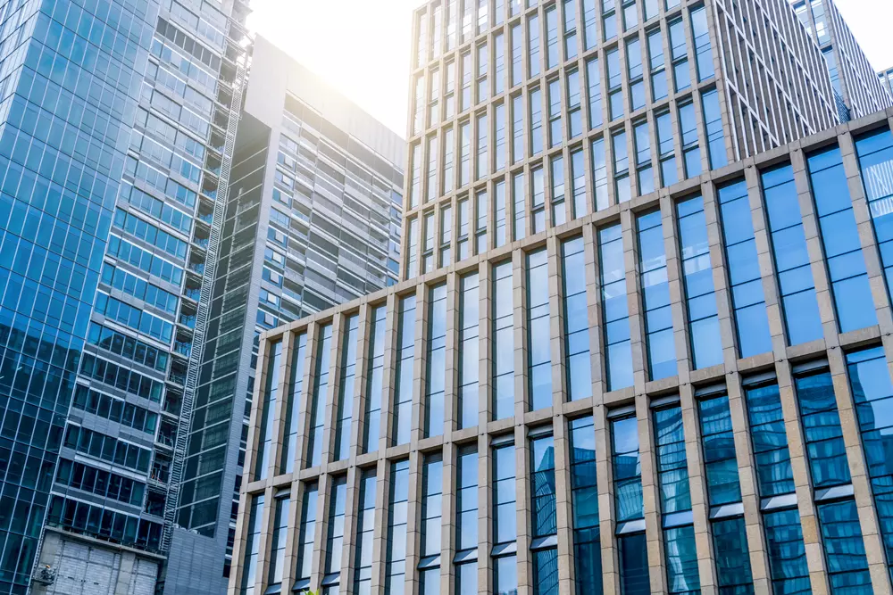 Modern skyscrapers with large glass windows reflect sunlight in an urban setting. The buildings, which house various businesses and services, feature a mix of geometric designs and varying heights.