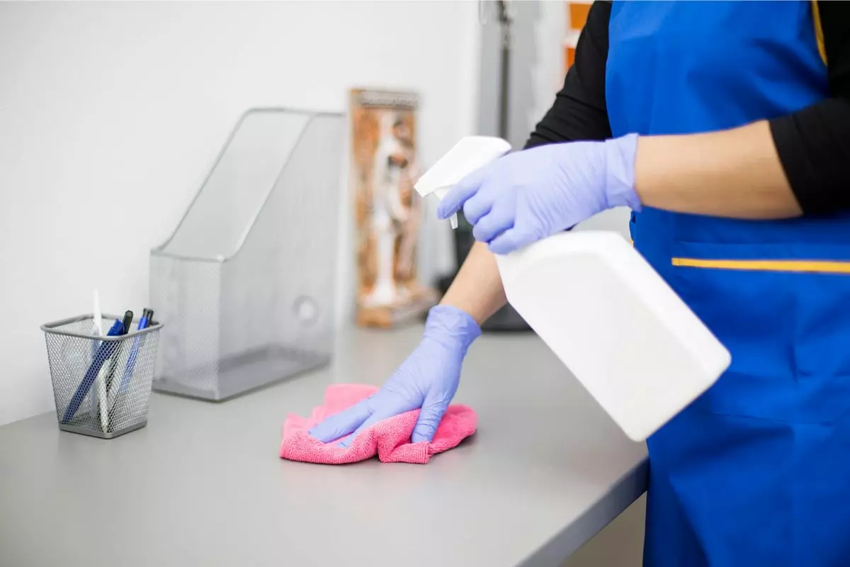 Person wearing blue apron and purple gloves uses a spray bottle and pink cloth to clean a gray desk, highlighting the meticulous services provided. A pen holder, file holder, and decorative item are on the desk.
