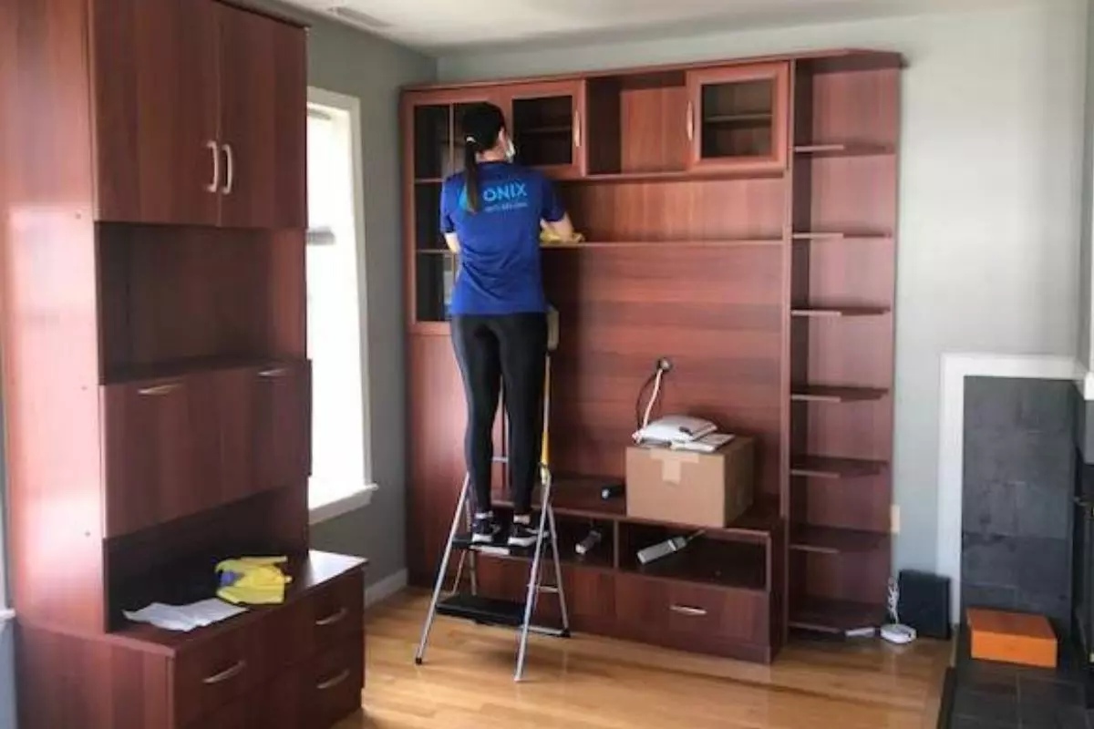 A person stands on a stepladder, organizing items on a tall wooden shelving unit in a room with hardwood floors, a window, and a fireplace. A cardboard box and various items are scattered around, hinting at recent home organization services.