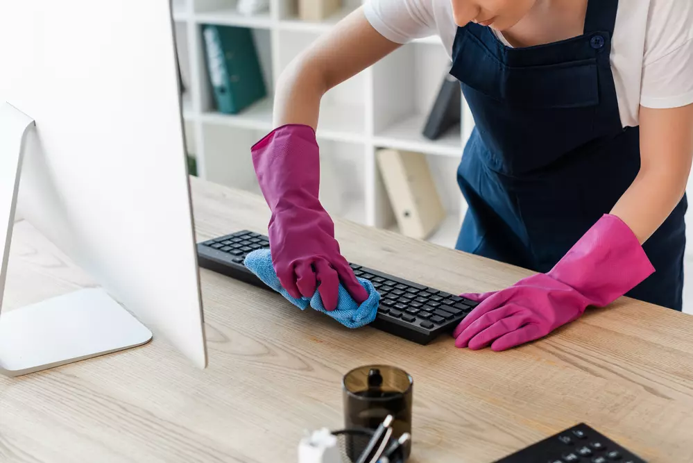 A person wearing pink gloves and a navy apron, diligently providing cleaning services, is seen wiping a keyboard with a blue cloth at a desk.