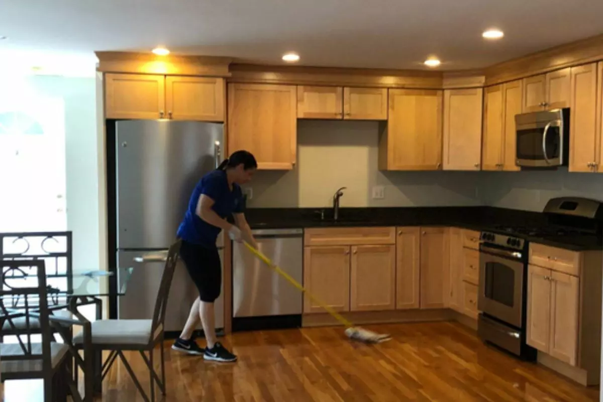 A person mops the wooden floor of a clean, modern kitchen with light wood cabinetry, stainless steel appliances, recessed lighting, showcasing professional cleaning services.