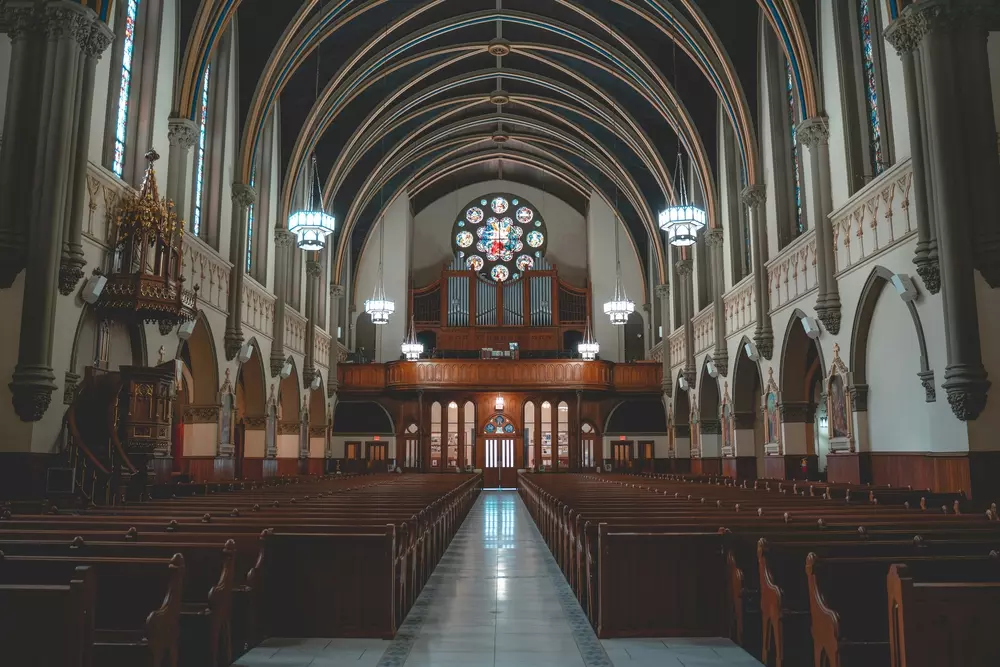 Interior view of a large, ornate church with tall arched ceilings, stained glass windows, wooden pews, and chandeliers, ready to host its timeless services.