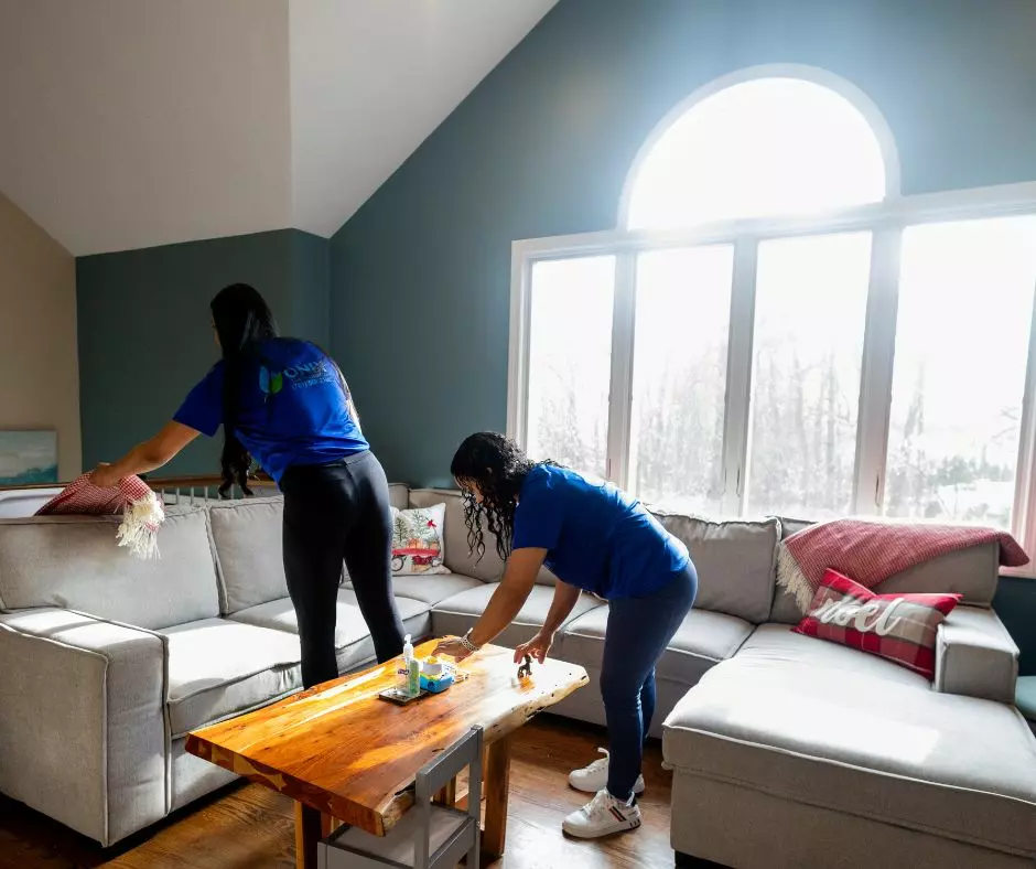 Two people in blue shirts methodically follow a cleaning checklist as they tidy a living room with a large sectional sofa, wooden coffee table, and a big window letting in natural light. One is adjusting a cushion while the other wipes down the table.