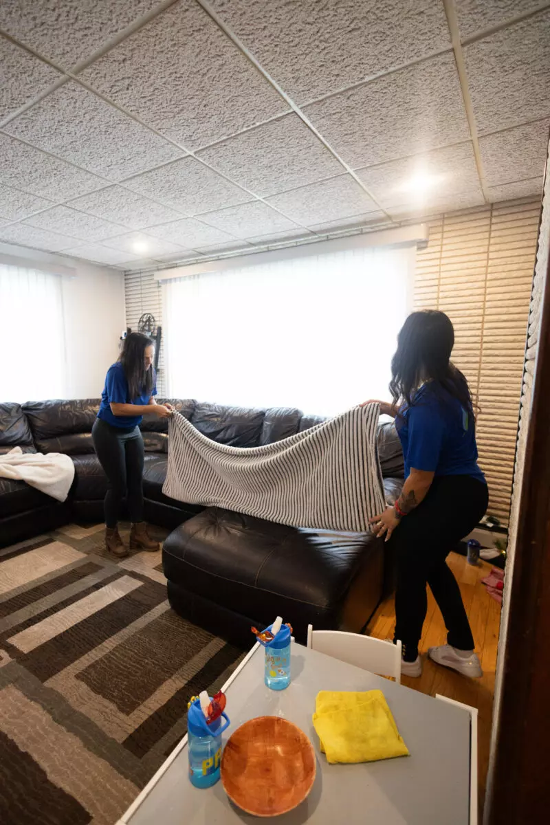 Two individuals are tidying up a living room, spreading a striped blanket on a black sectional sofa. Cleaning supplies and a bowl are neatly arranged on a nearby table, demonstrating their commitment to quality services.