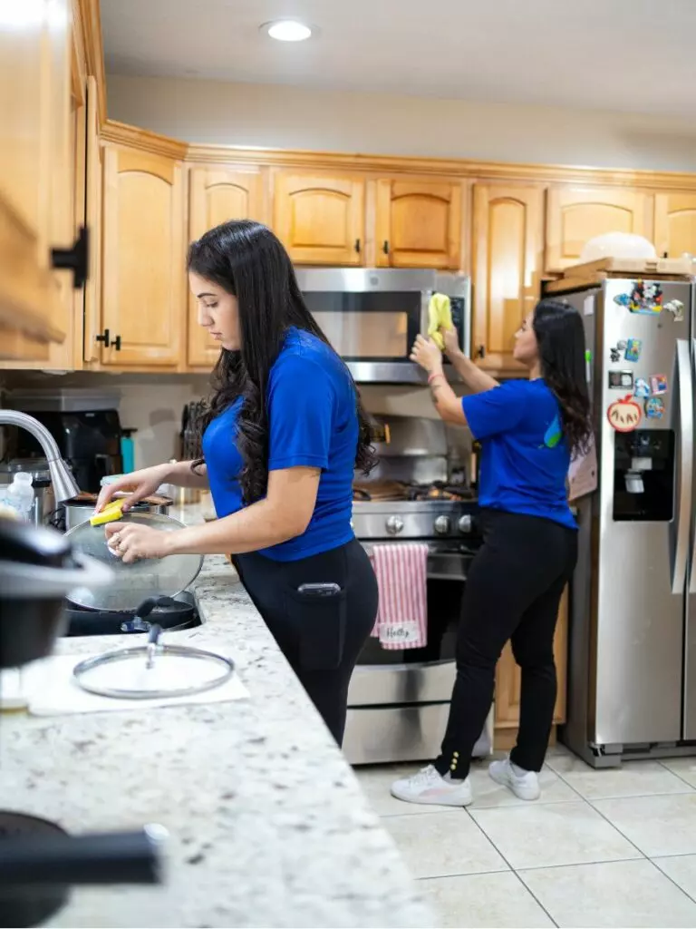 Two women in blue shirts clean a kitchen as meticulously as curators arranging an art gallery. One wipes down a stovetop while the other cleans a microwave. The kitchen, with its wooden cabinets and stainless steel refrigerator, gleams like an exhibition space.