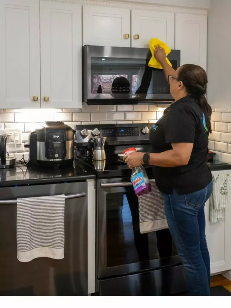 In a modern kitchen, a person meticulously cleans a microwave with a yellow cloth, while holding a spray bottle, creating a scene that could belong in an art gallery.