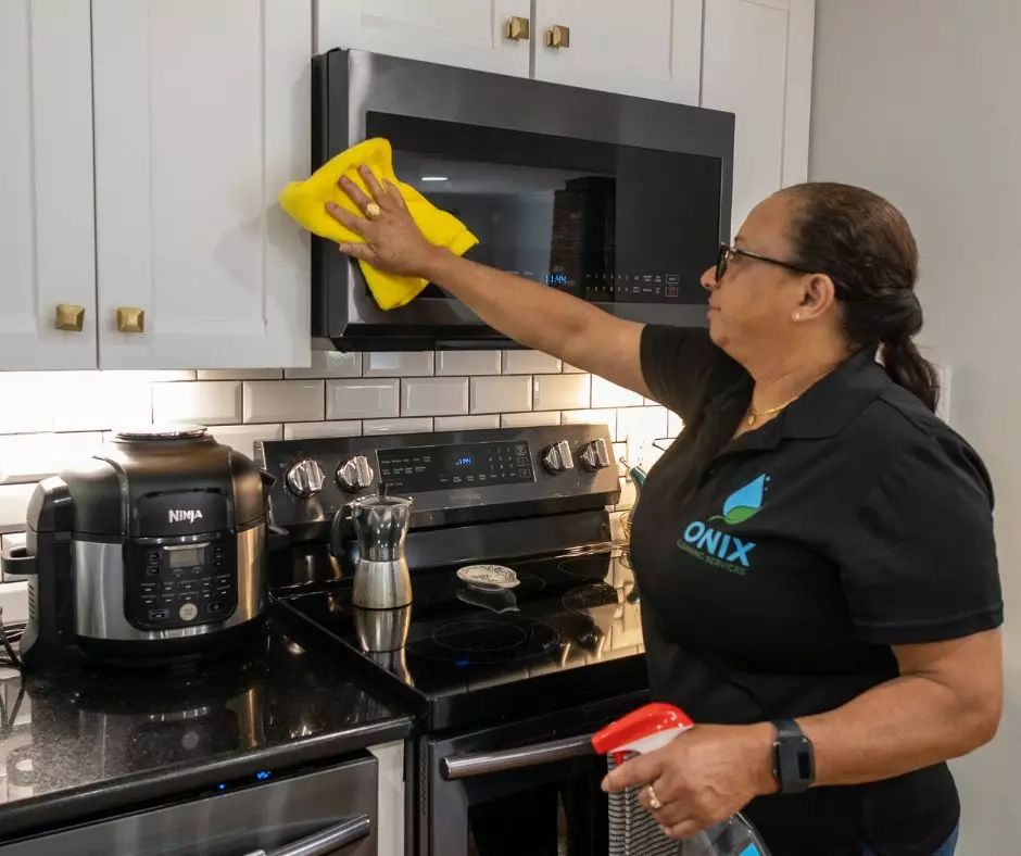 A person cleaning a microwave in a kitchen, using a yellow cloth. They are wearing a black shirt with an "Onix" logo, resembling an exhibit in a modern gallery. A pressure cooker and cleaning spray are on the countertop.