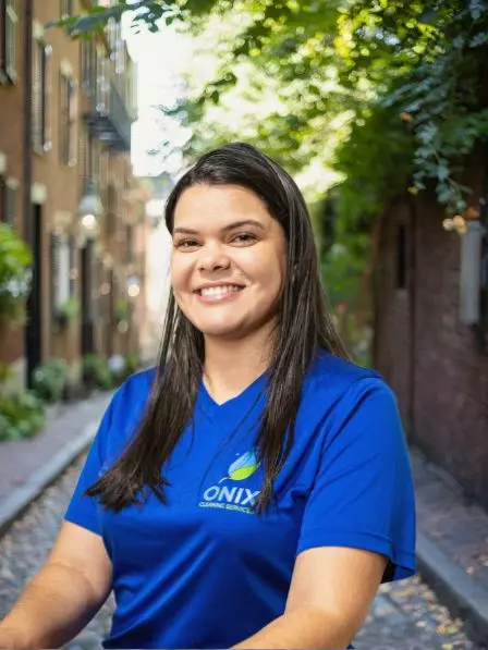 A woman with long dark hair wearing a blue shirt labeled "Onix Cleaning Services" smiles while standing on a cobblestone street lined with brick buildings and trees.