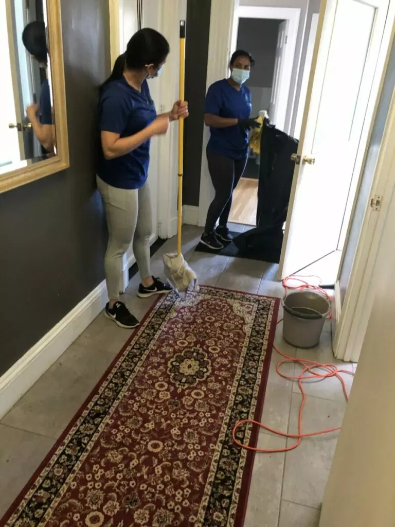 Two individuals in blue shirts, one mopping a hallway floor and the other holding a trash bag, appear to be preparing an exhibition space. A red patterned rug, gray mop bucket, and an orange cord are visible in the scene.