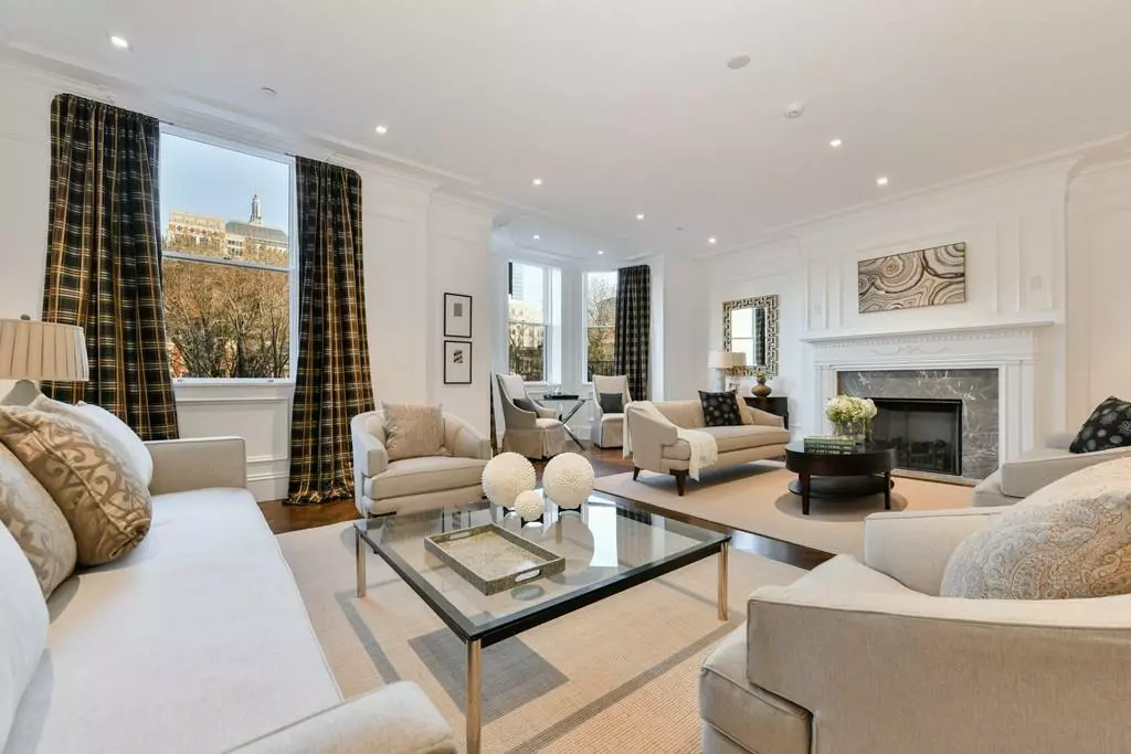 Spacious, elegant living room with a gallery-like arrangement featuring multiple seating areas, a fireplace, large windows with plaid curtains, and a glass coffee table. The room has neutral-colored furniture and a light, airy feel.