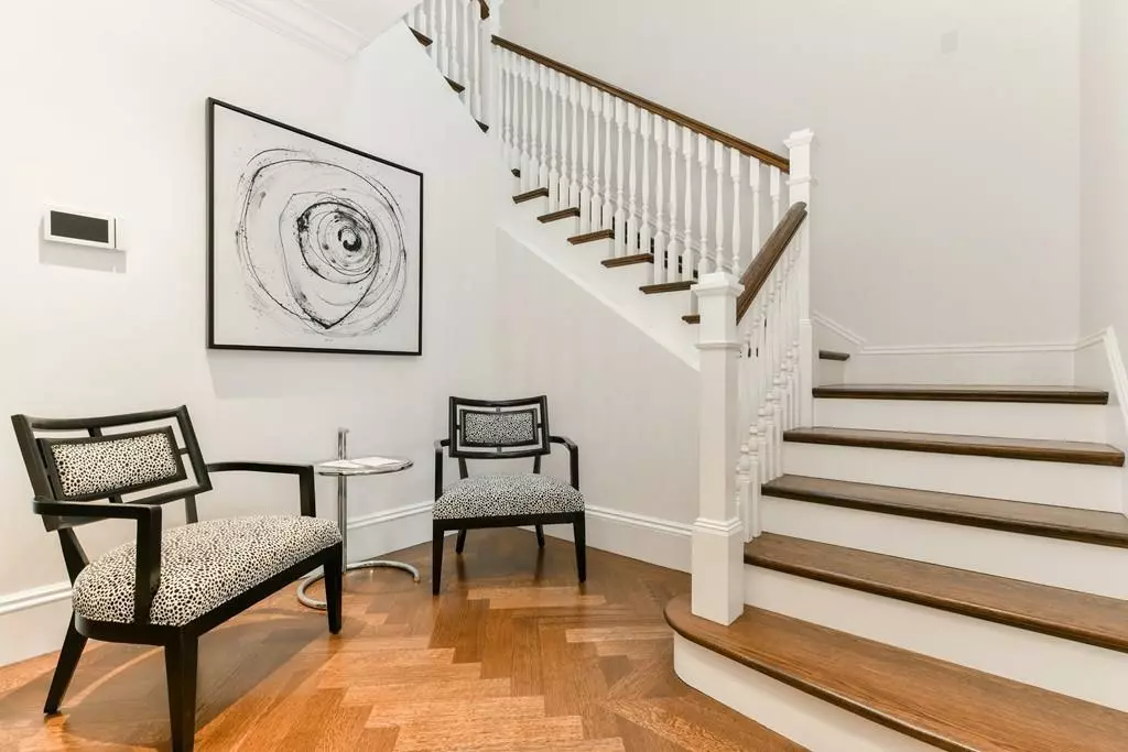 A modern hallway with wooden stairs, two patterned chairs, a small round table, and a black-and-white abstract painting on the wall, evoking a gallery-like ambiance.