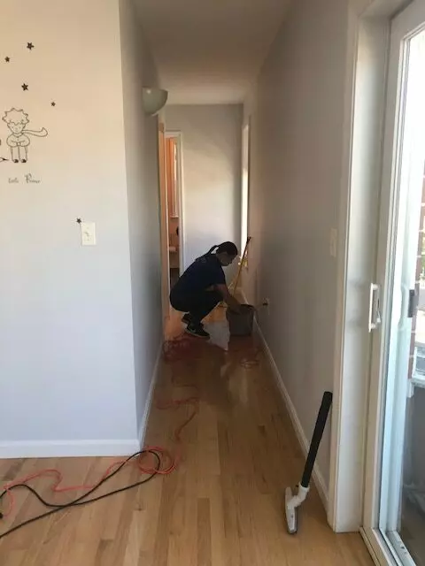 A person mops the hallway floor using a bucket and mop, with visible extension cords snaking across the tiles. A wall decal adds a touch of charm, while the hallway leads to a brightly lit gallery room at the end.