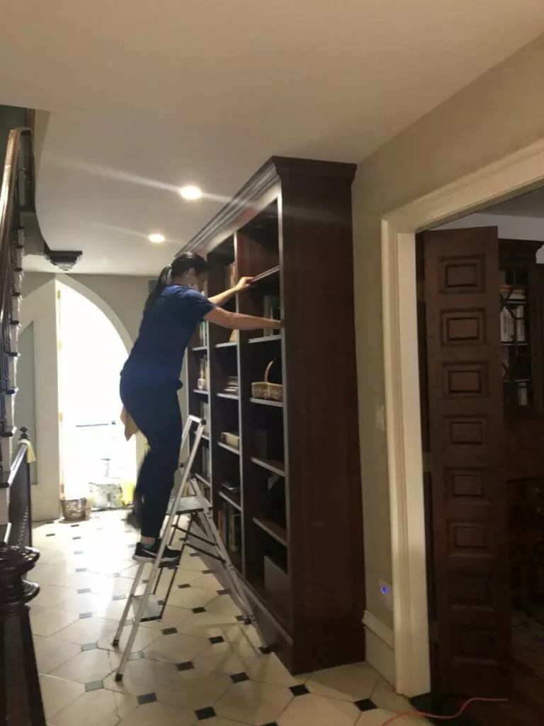 A person on a ladder reaching up to organize shelves on a tall wooden bookcase in a well-lit gallery with tiled flooring and an open door.