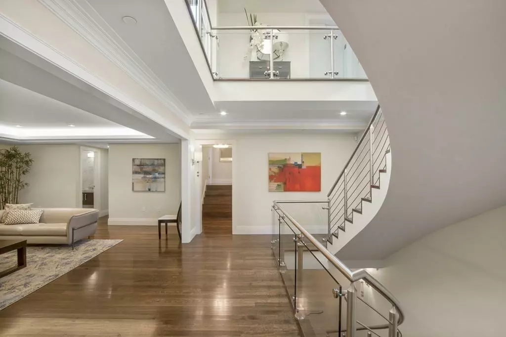 Modern, open-plan living area with wooden flooring, an elegant staircase with glass railings, and contemporary artwork on the walls that evokes a gallery feel. A beige sofa is positioned near a small table.