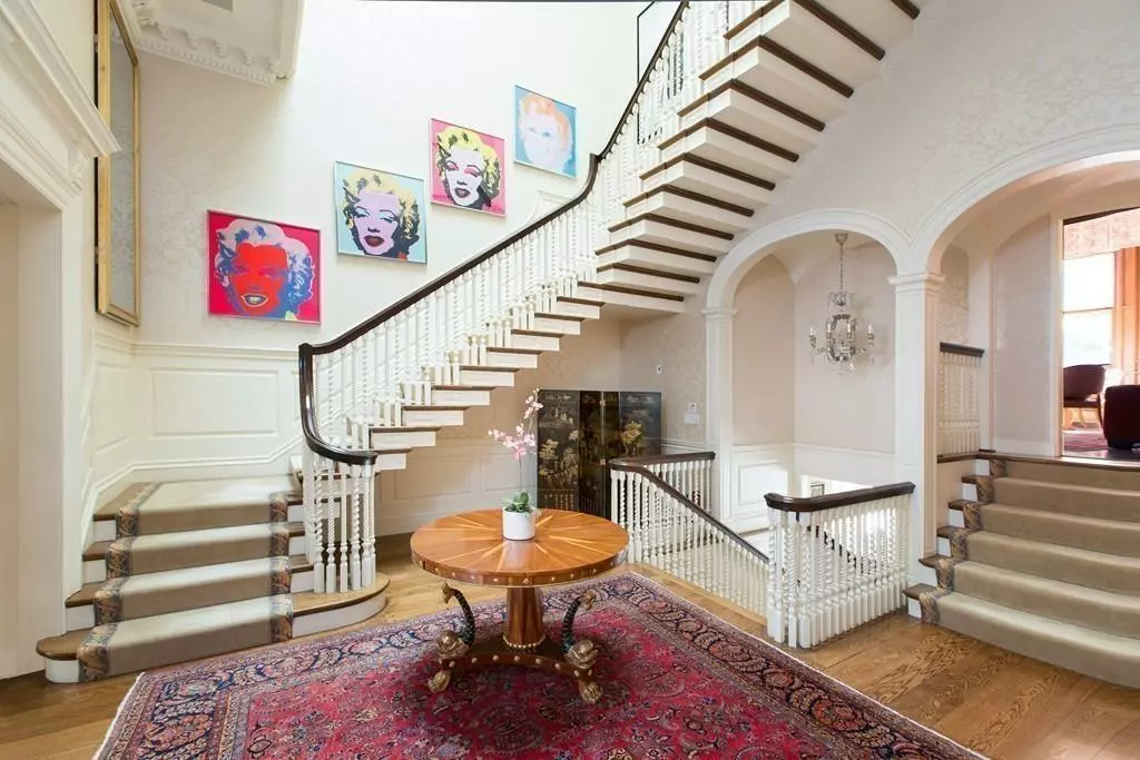 A grand staircase with wooden steps and white railings graces a well-lit room with white walls. A round wooden table with a potted plant sits on a red patterned rug, while four colorful pop art portraits are displayed on the wall, creating an inviting gallery-like atmosphere.