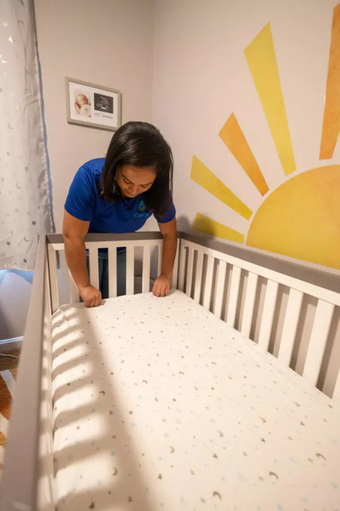 A person in a blue shirt is adjusting a sheet in a white crib. The wall behind has a yellow sun mural, reminiscent of an art gallery exhibition, and there are pictures hanging on the adjacent wall.