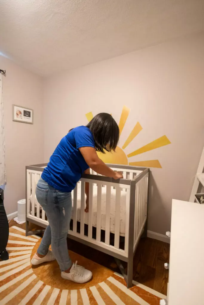 A woman leans over a baby crib with a sun mural on the wall in a nursery that resembles an art gallery. She is wearing a blue shirt and jeans, and the room has a warm color scheme.