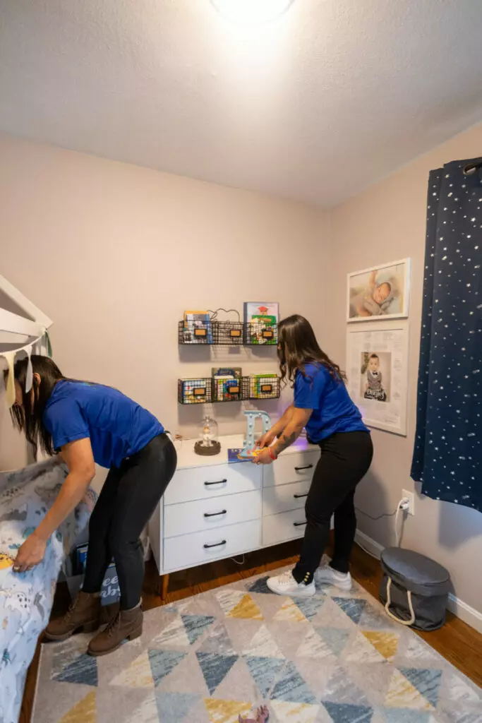 Two individuals in blue shirts are tidying a child's room, one on the left arranging items on the bed and the other on the right organizing objects on a white dresser, making the space look like a neat little gallery.