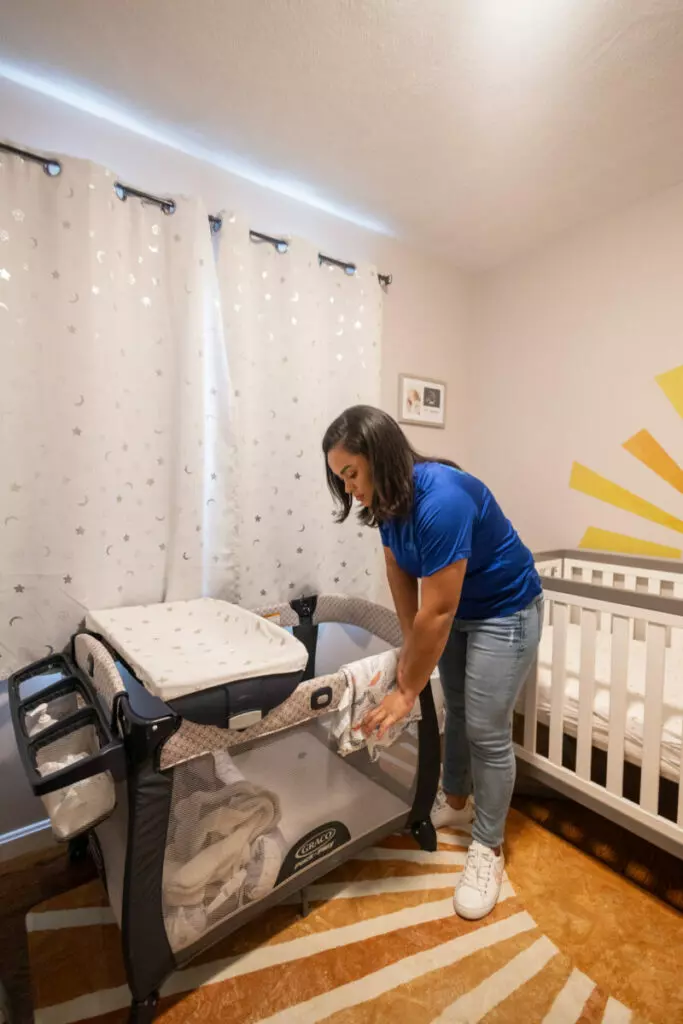 A person in a blue shirt is arranging a blanket in a baby crib situated in a nursery room with white curtains and a yellow sun wall decoration, making the space feel like an intimate art gallery.