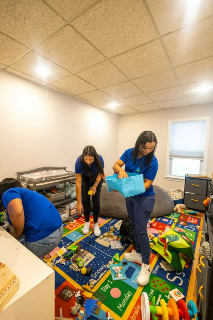 Three people are cleaning and organizing a brightly colored children's playroom, which resembles a lively gallery with toys scattered on the floor.