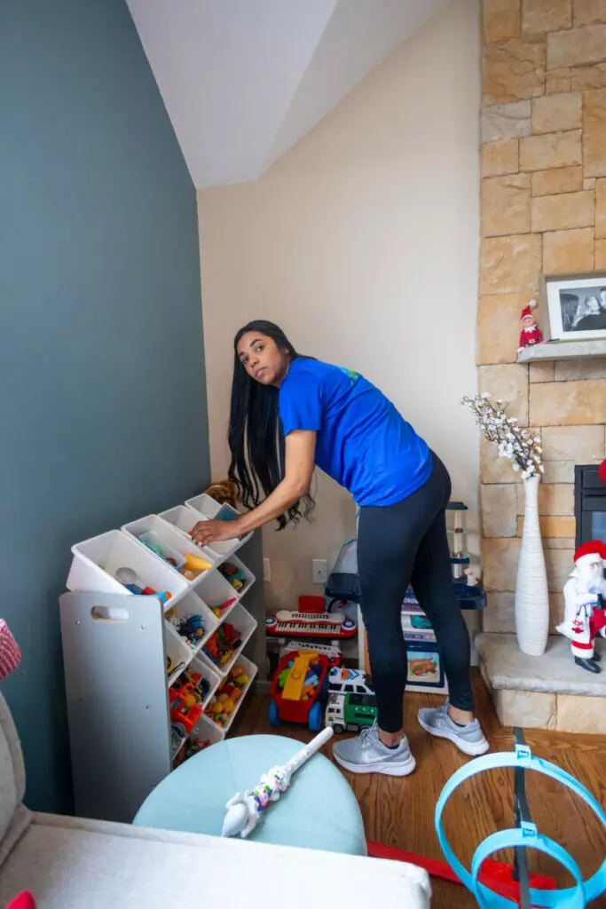 A person is organizing toys in a white shelf next to a stone fireplace, creating a gallery of playful memories. A vacuum cleaner and various toys are scattered on the floor. The room has green and beige walls.