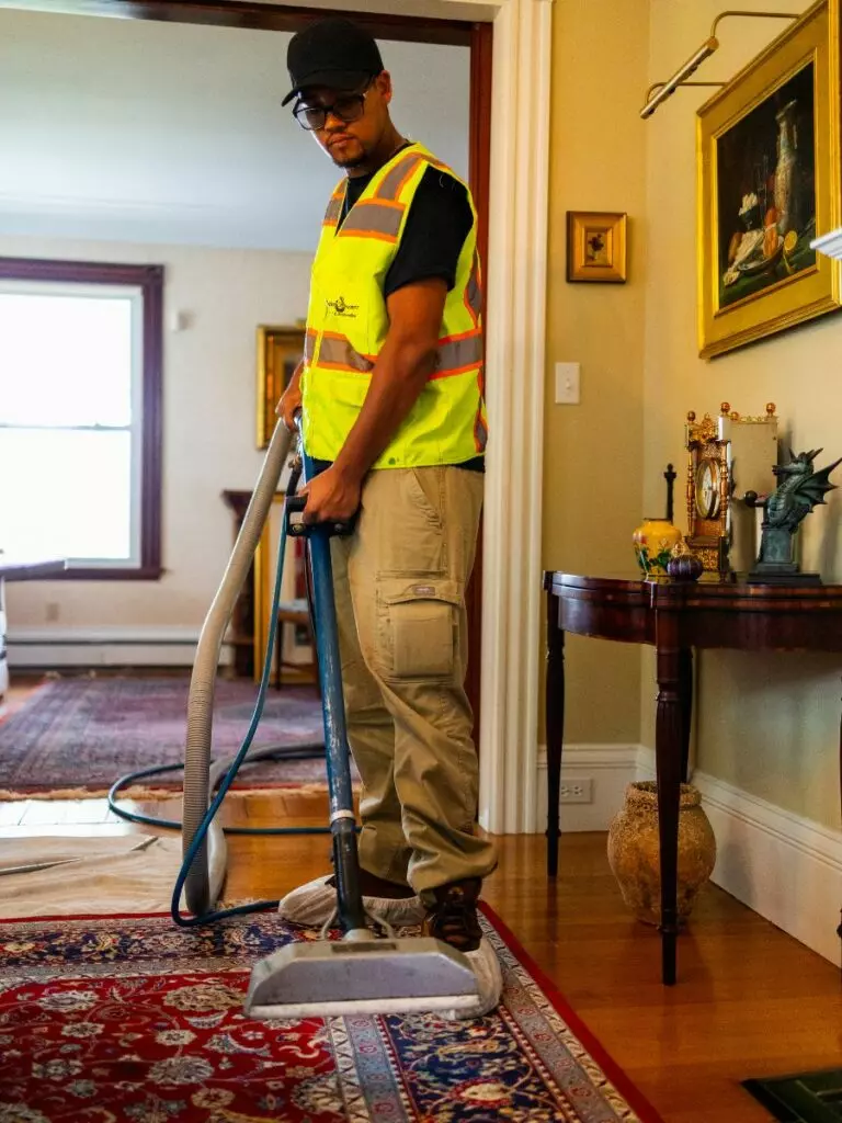 A man in a high-visibility vest and cap performs interior maintenance by using a vacuum to clean a red patterned rug in a well-decorated room.