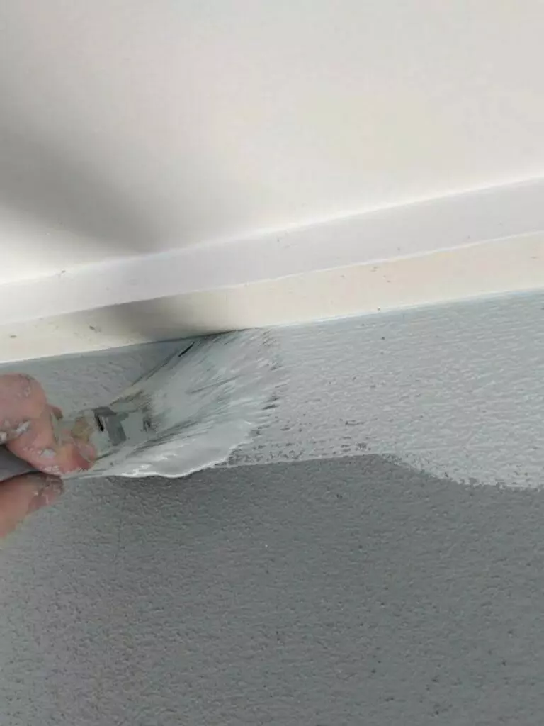 A hand is using a paintbrush to apply white paint along the edge of a wall, overlapping onto the grey surface below—a meticulous task in interior maintenance.