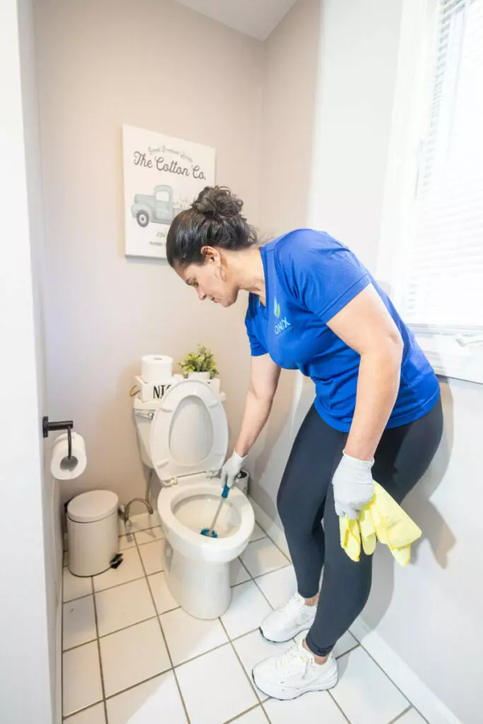 A woman in a blue shirt and black leggings cleans a white toilet with a brush in a small, bright bathroom.