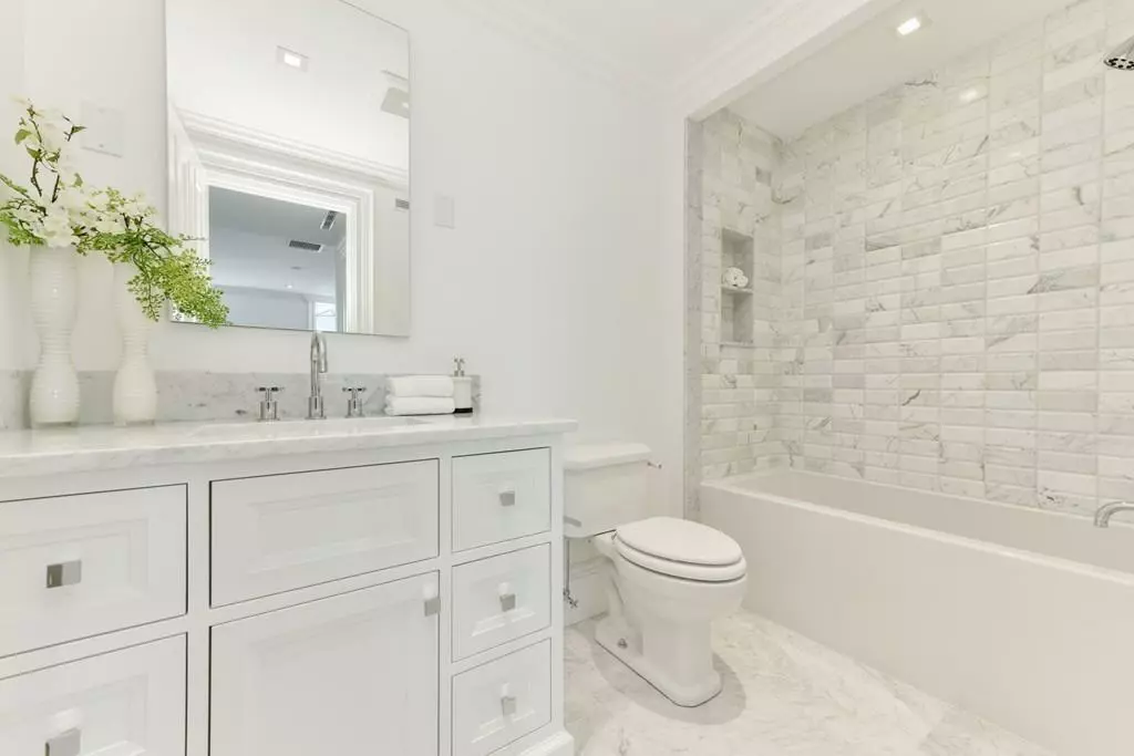 A white bathroom with a vanity, mirror, toilet, and bathtab. The bathtub area is decorated with grey marble tiles. White vases with flowers are on the countertop.