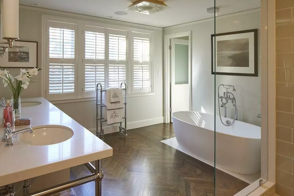 Bright bathroom with wooden parquet flooring, a freestanding bathtub, a double sink vanity, wall-mounted towel rack, framed wall art, and windows with white shutters.